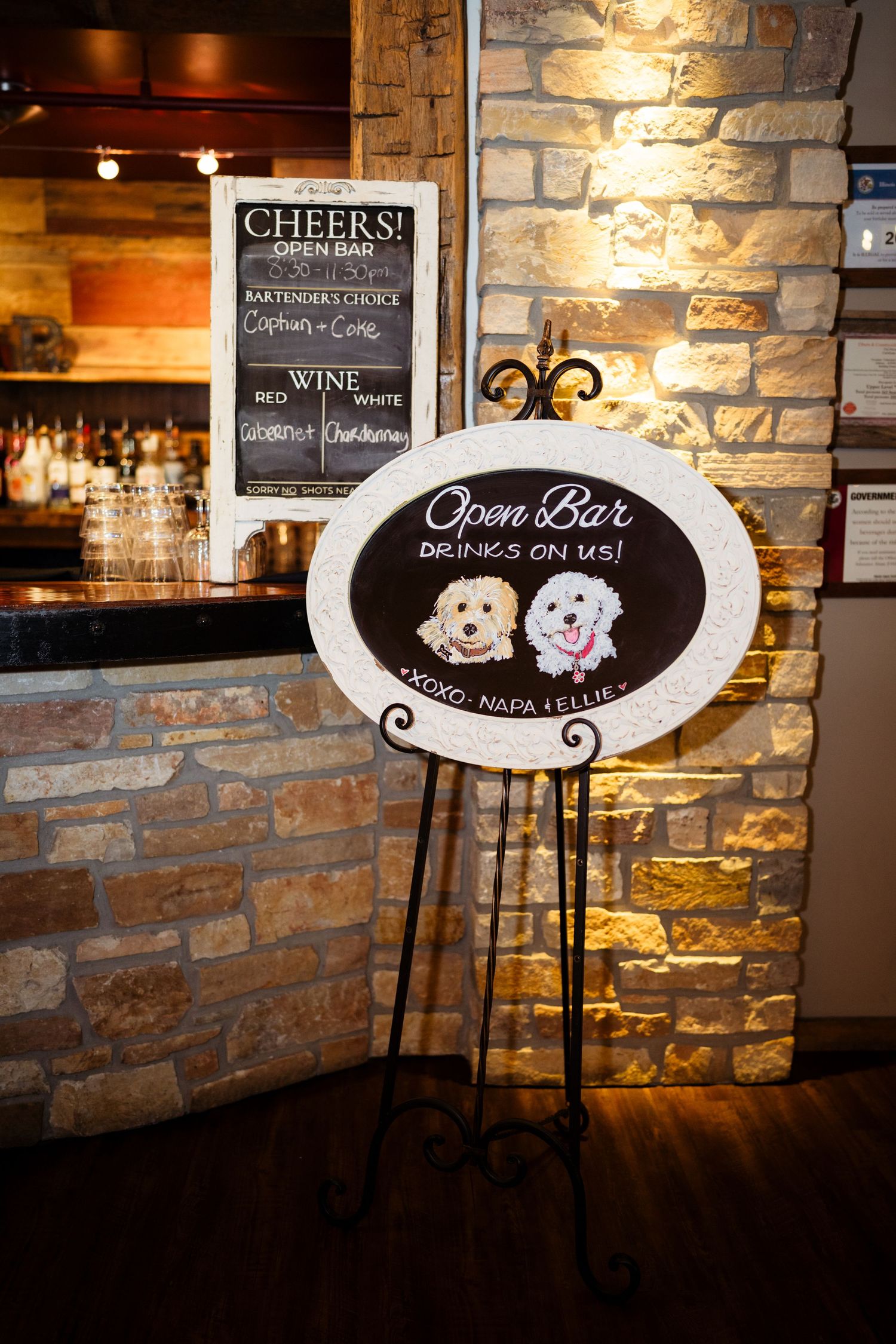 Open bar chalkboard sign on easel reads Drinks On Us with cute dog illustrations at rustic stone wedding venue.