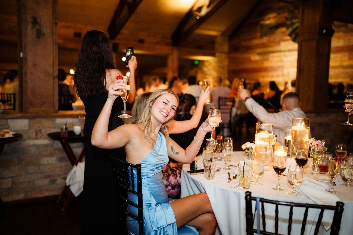 Guests celebrate at elegant wedding reception, raising wine glasses and laughing at candlelit tables in rustic venue.