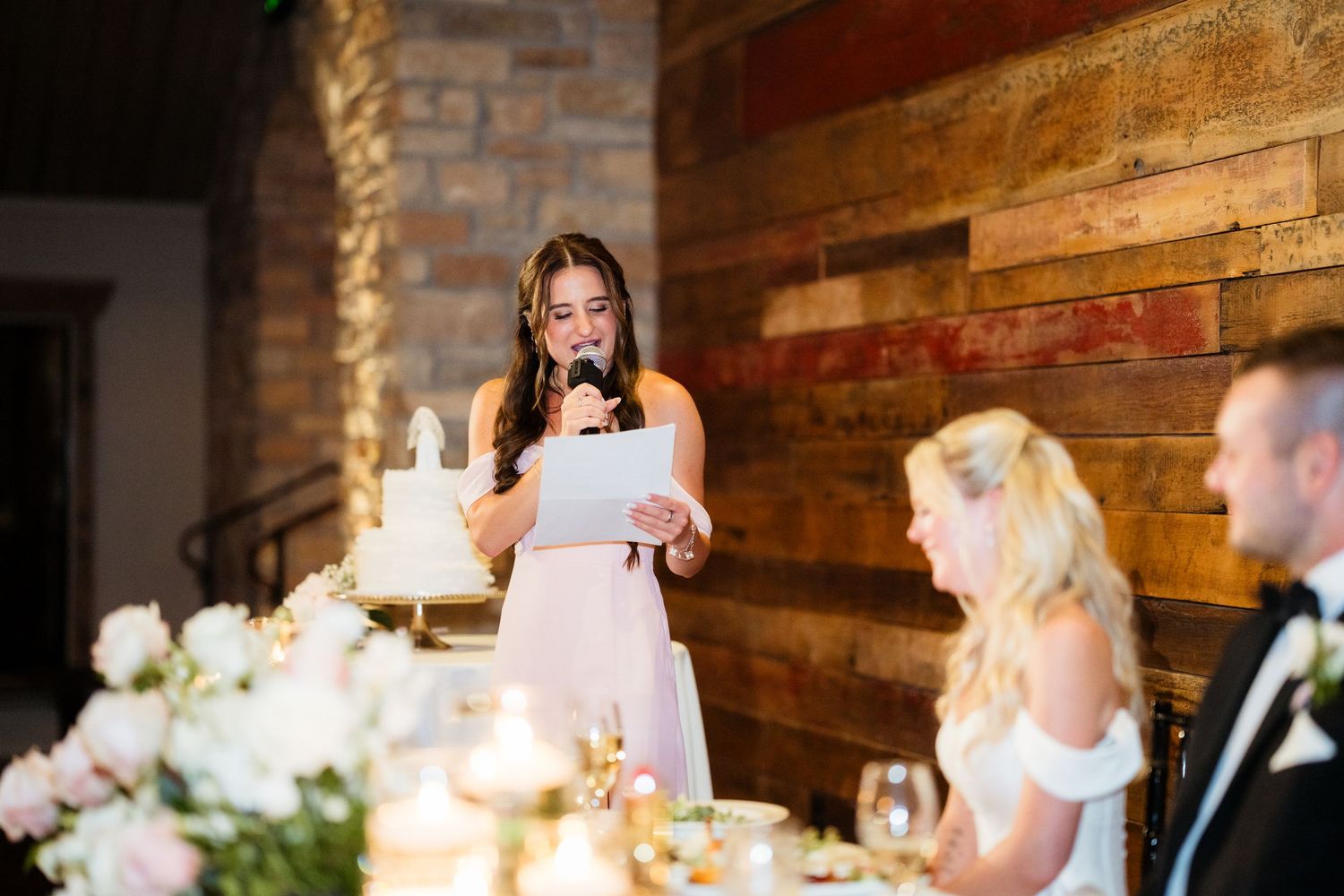 Bridesmaid giving a heartfelt speech at wedding reception with rustic wood wall backdrop and floral centerpieces.