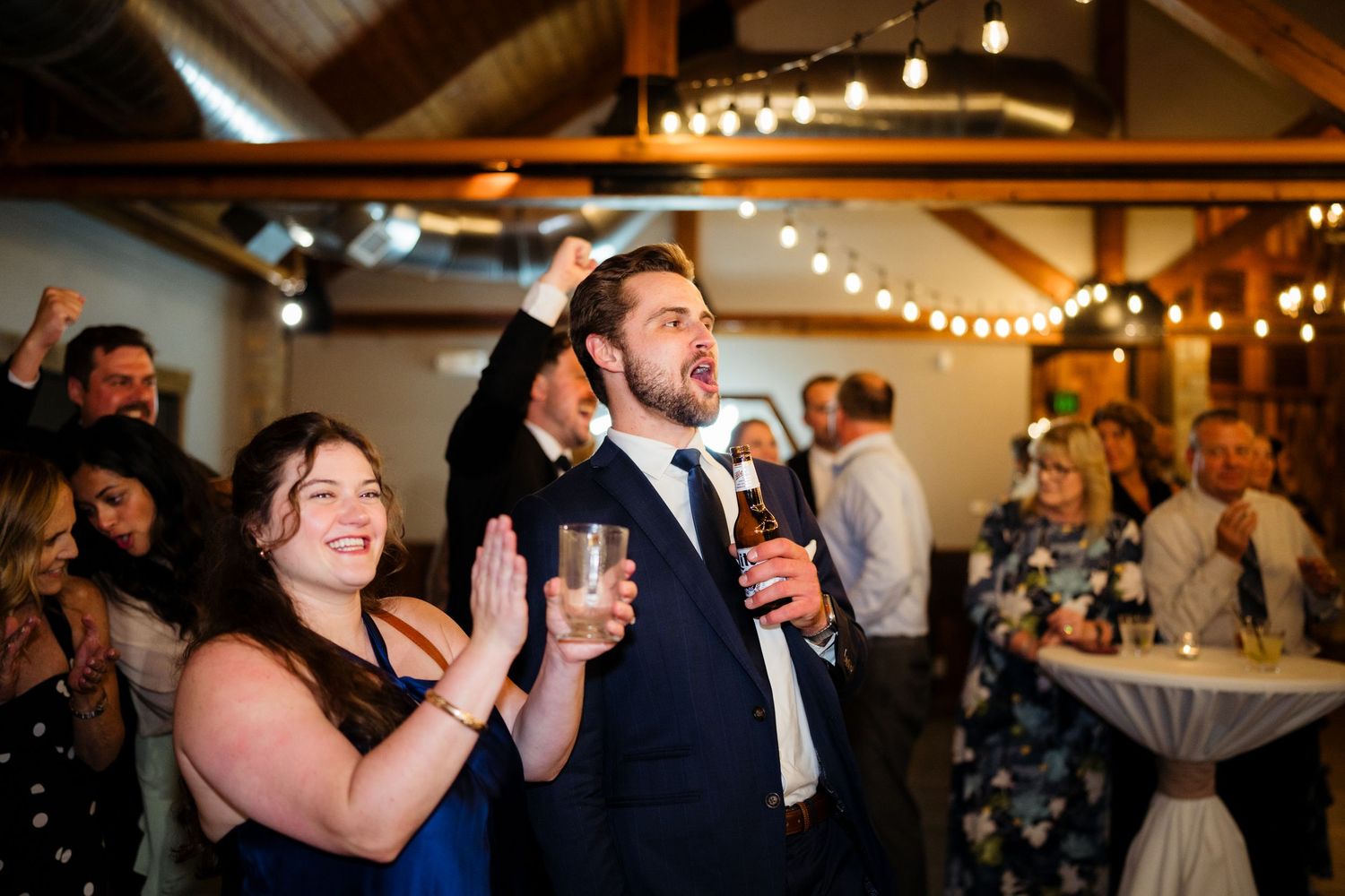 Wedding guests clapping and cheering at a reception venue decorated with string lights.
