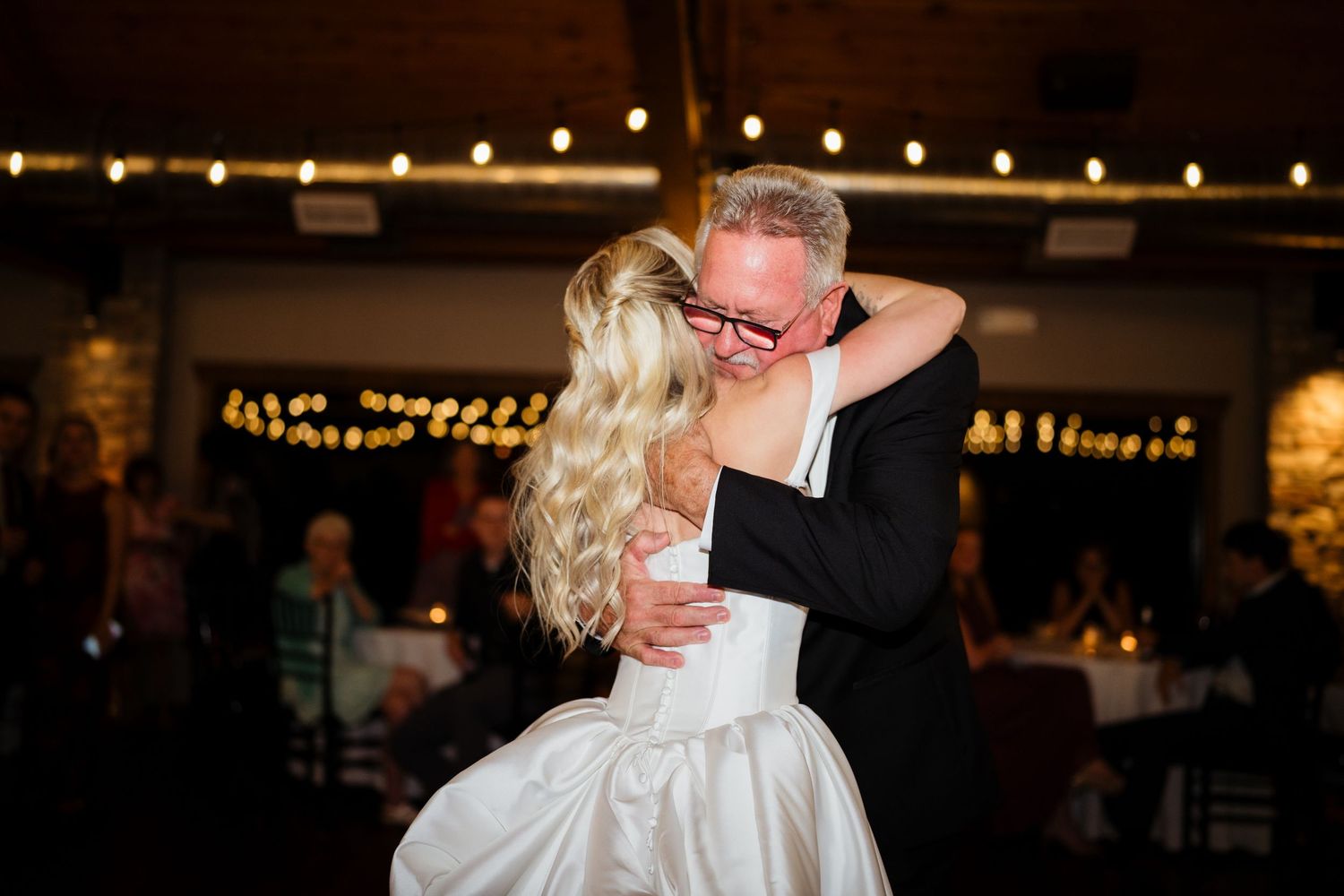 Bride and father share an emotional first dance hug at a warmly lit wedding reception venue.