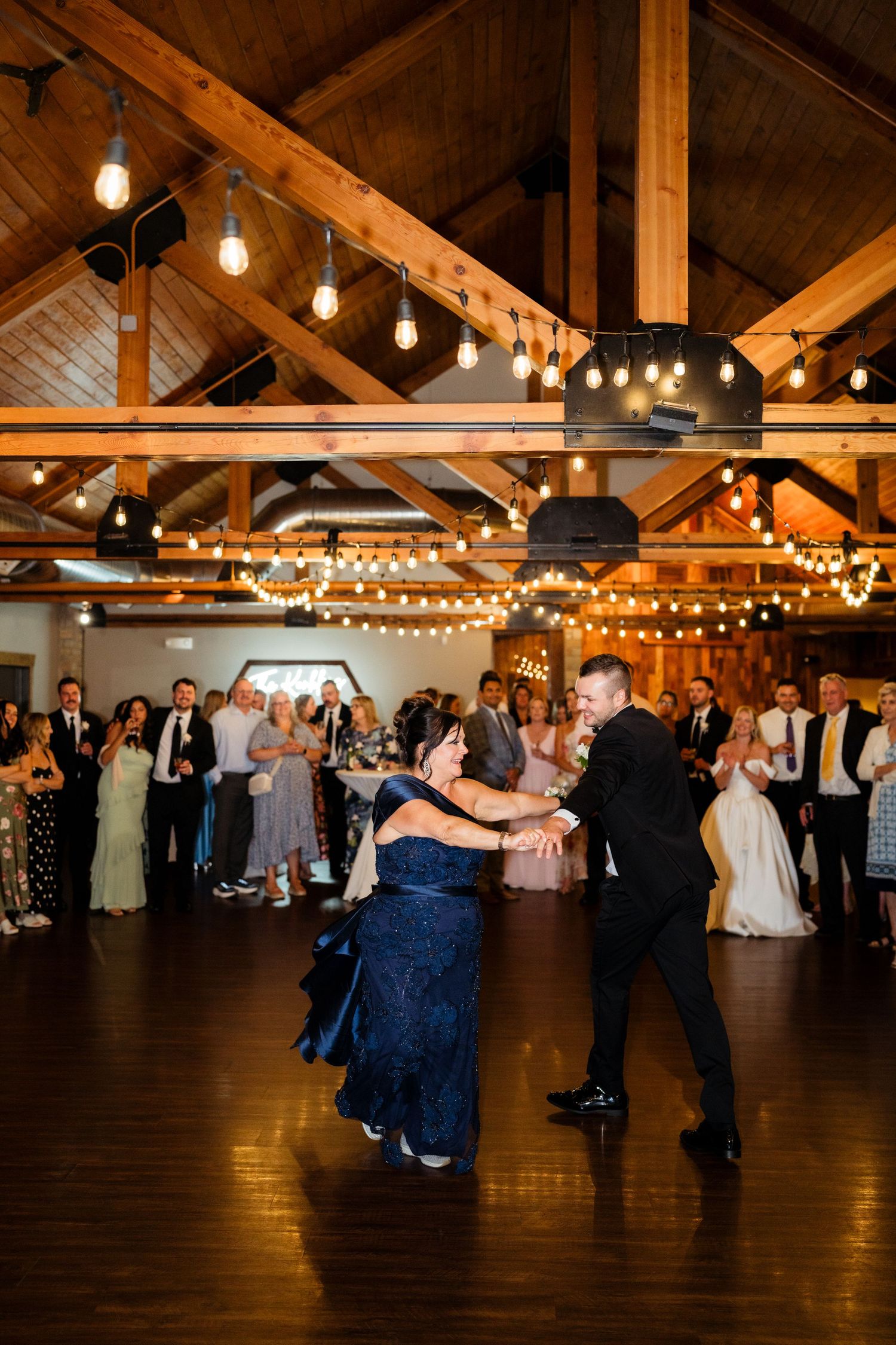 Groom dances with mother in elegant barn wedding venue with string lights and exposed wood beams.