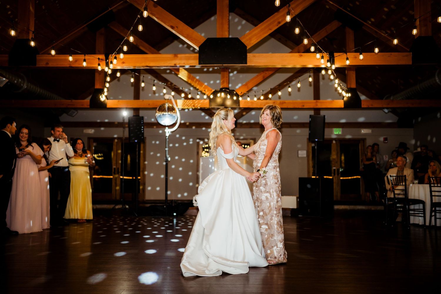 Two brides share their first dance in a warmly lit barn wedding venue adorned with string lights and wooden beams.