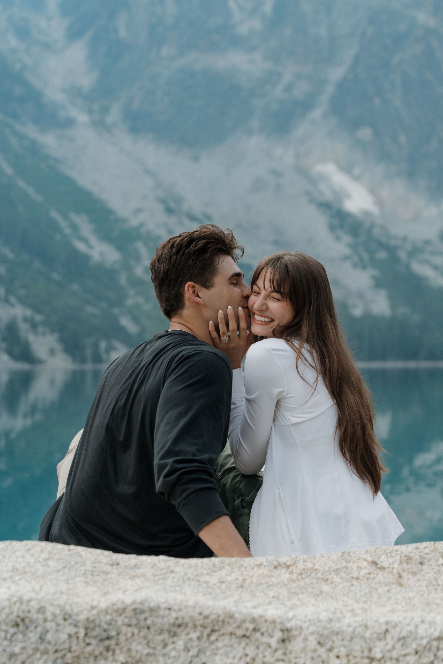 Couple sharing a romantic moment on a rocky ledge with a stunning mountain lake backdrop.