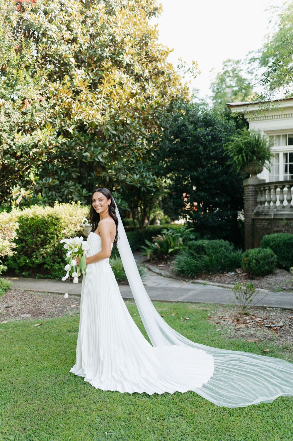 Bride in white pleated gown with long cathedral veil holding bouquet in lush garden.