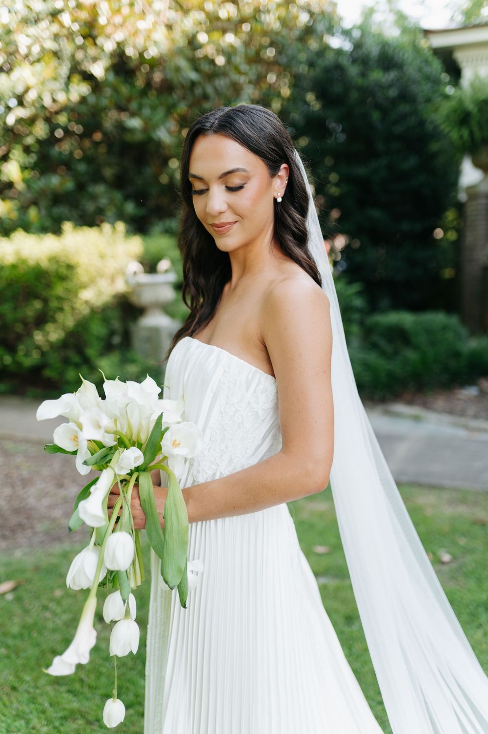 Bride in strapless white pleated gown holding cascading white tulip bouquet with veil, posing outdoors in lush garden.