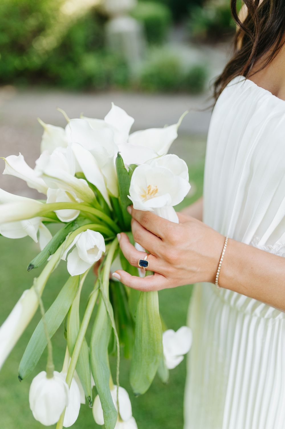 Bride in white dress holding elegant bouquet of white tulips and calla lilies, wearing sapphire ring and gold bracelet.