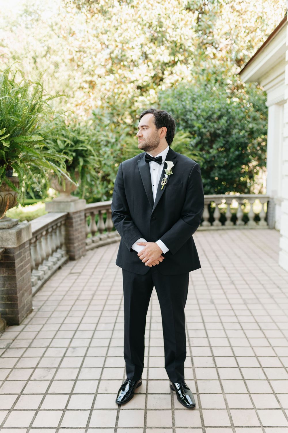 Dapper groom in classic black tuxedo with boutonniere standing on elegant stone terrace surrounded by lush greenery.