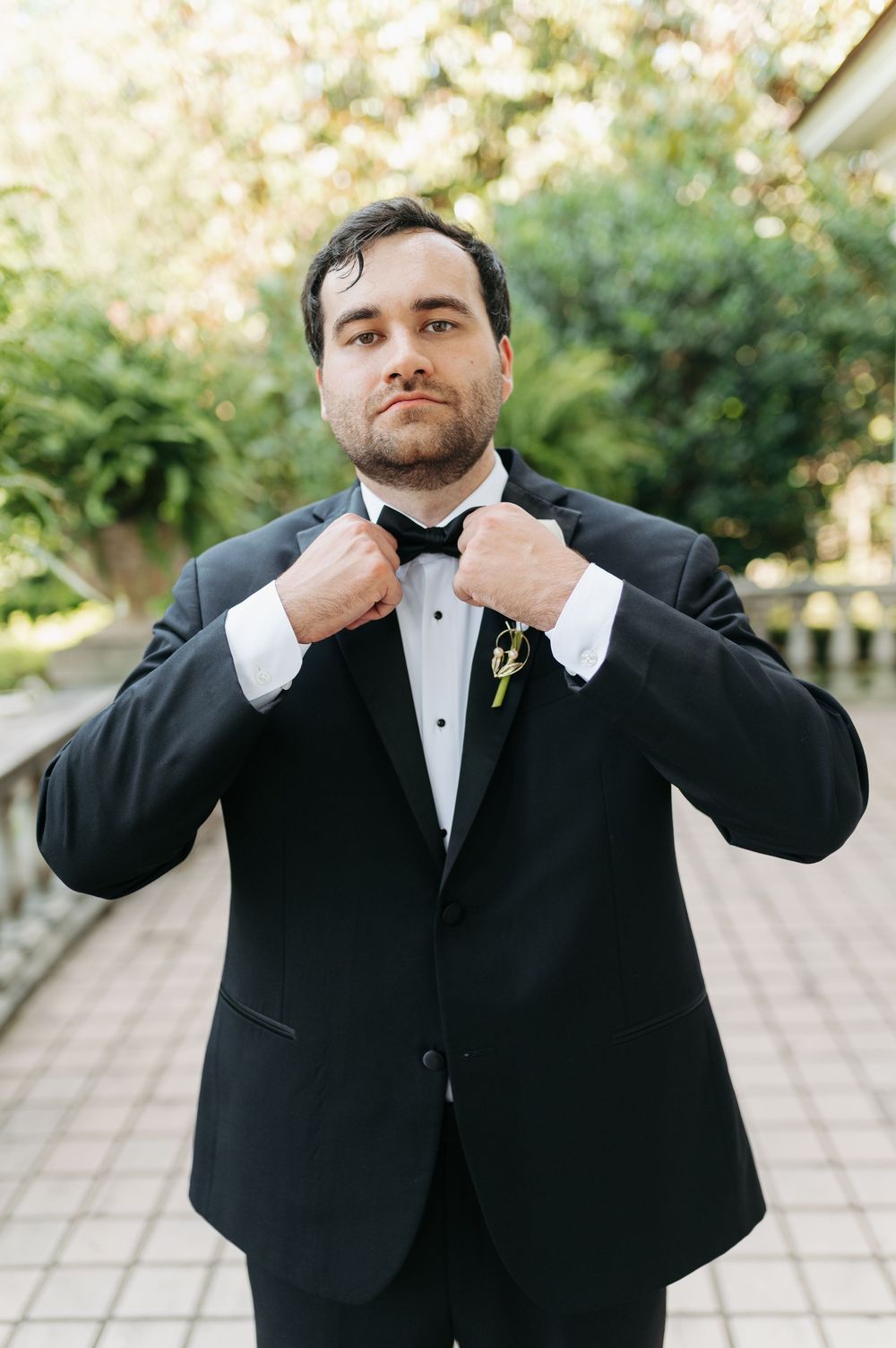 Groom in black tuxedo adjusting bow tie outdoors, wearing boutonniere, with garden balustrade in background.