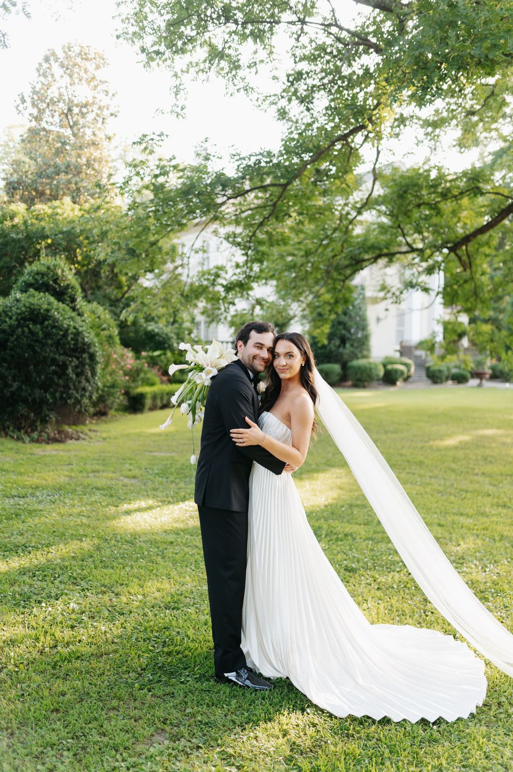 Bride in white pleated gown with veil embraces groom in black tuxedo outdoors on lush green lawn.