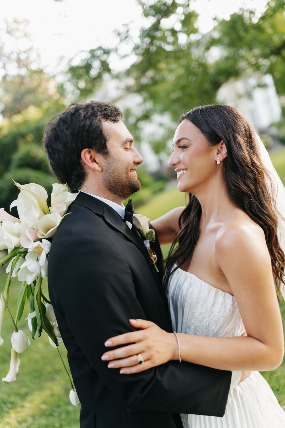 A smiling bride in a strapless gown embraces a bearded groom in a black tuxedo outdoors, holding white calla lilies.