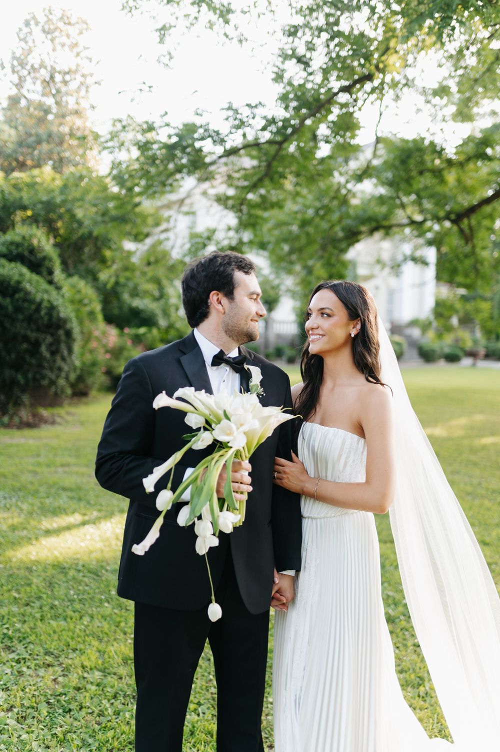 Smiling bride in strapless pleated white gown with veil and groom in black tuxedo holding white calla lily bouquet outdoors.