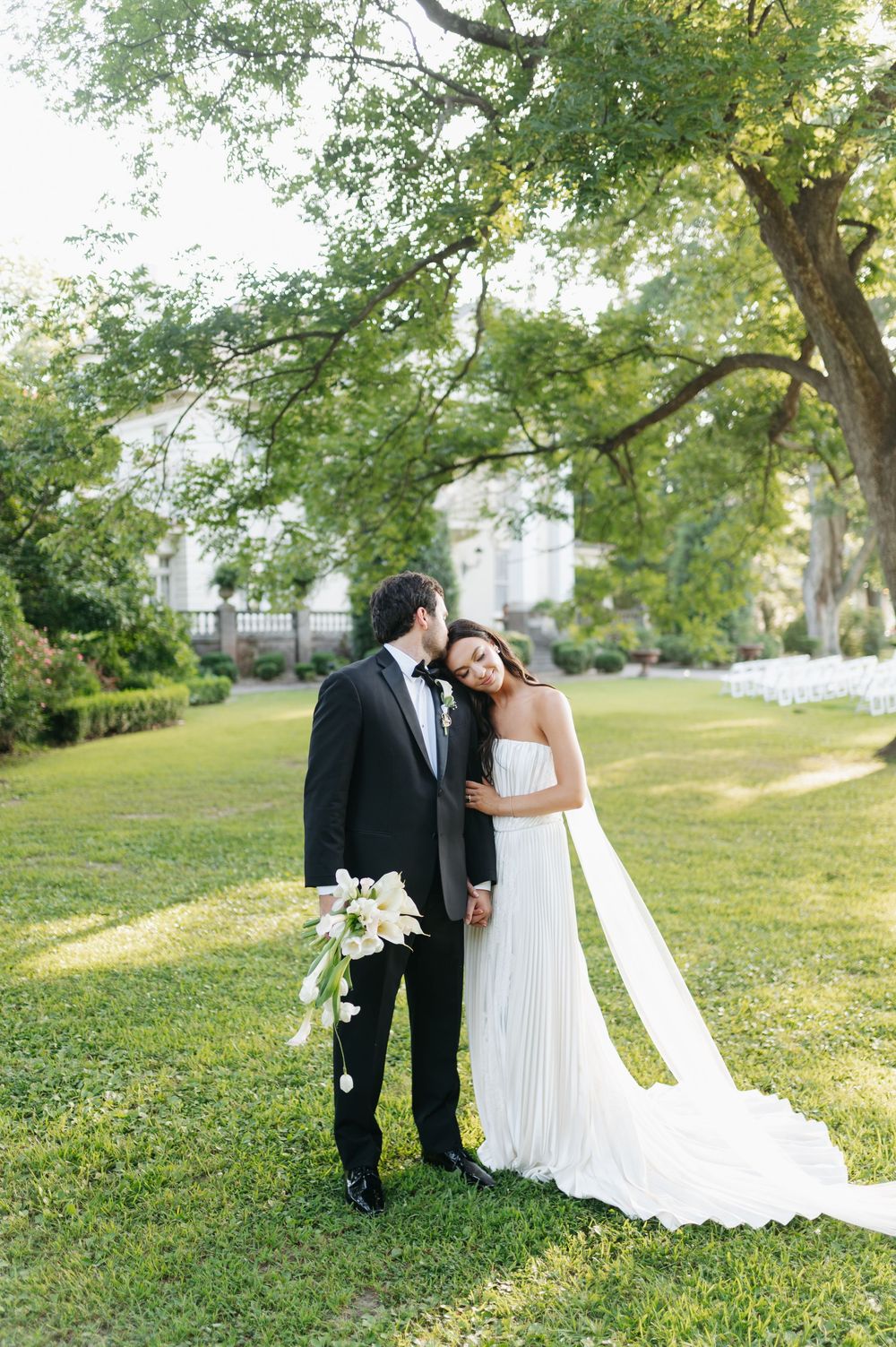 Bride and groom embracing on green lawn, she in white pleated gown with veil, he in black tuxedo holding calla lilies.