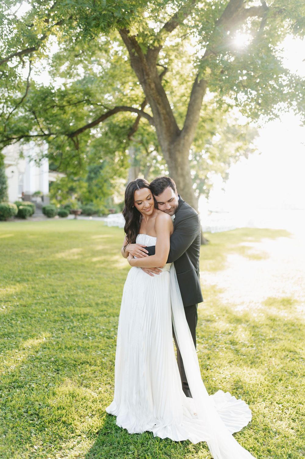 A bride and groom embrace tenderly under a large green tree on a sunny day, the bride wearing a flowing white strapless gown.