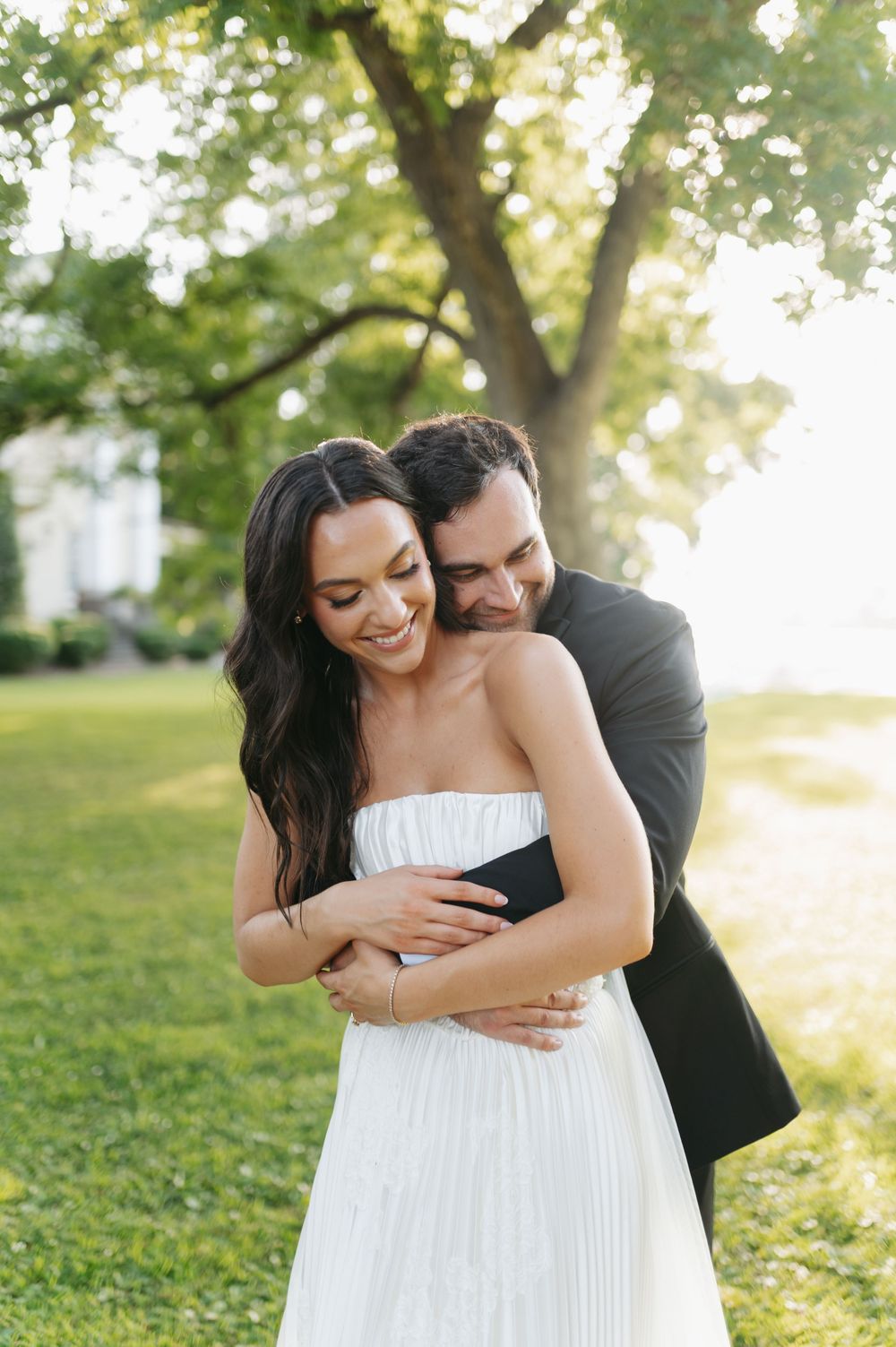 Groom in black tuxedo embracing smiling bride in strapless white dress outdoors.