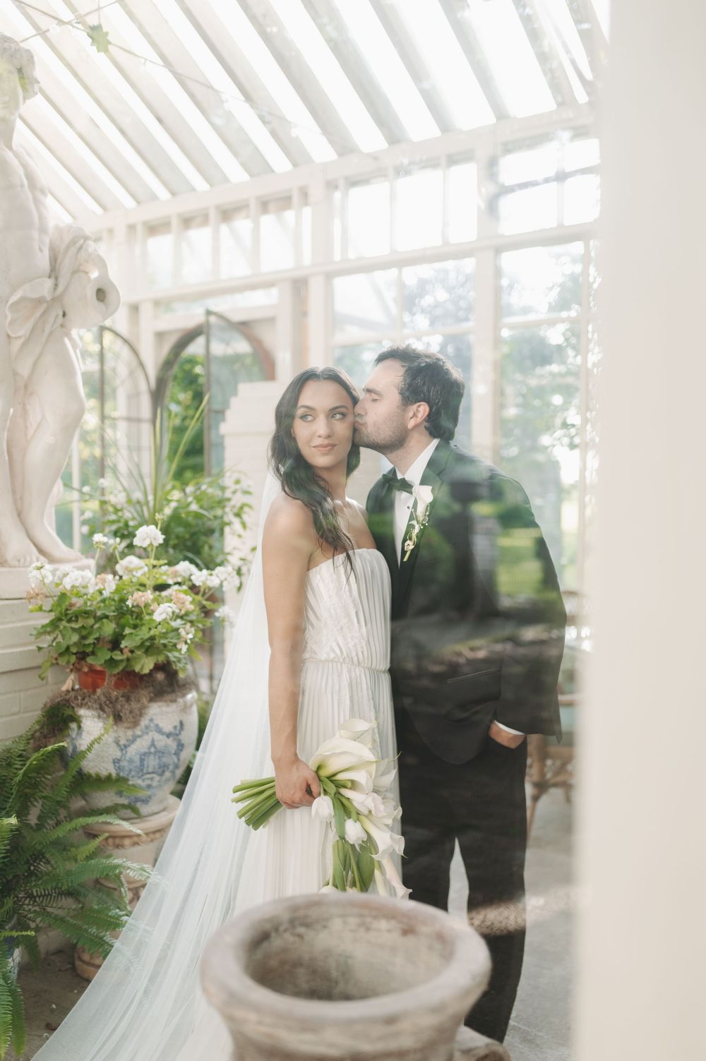 Bride and groom share a tender kiss in a sunlit greenhouse, bride holding white tulips in elegant wedding attire.