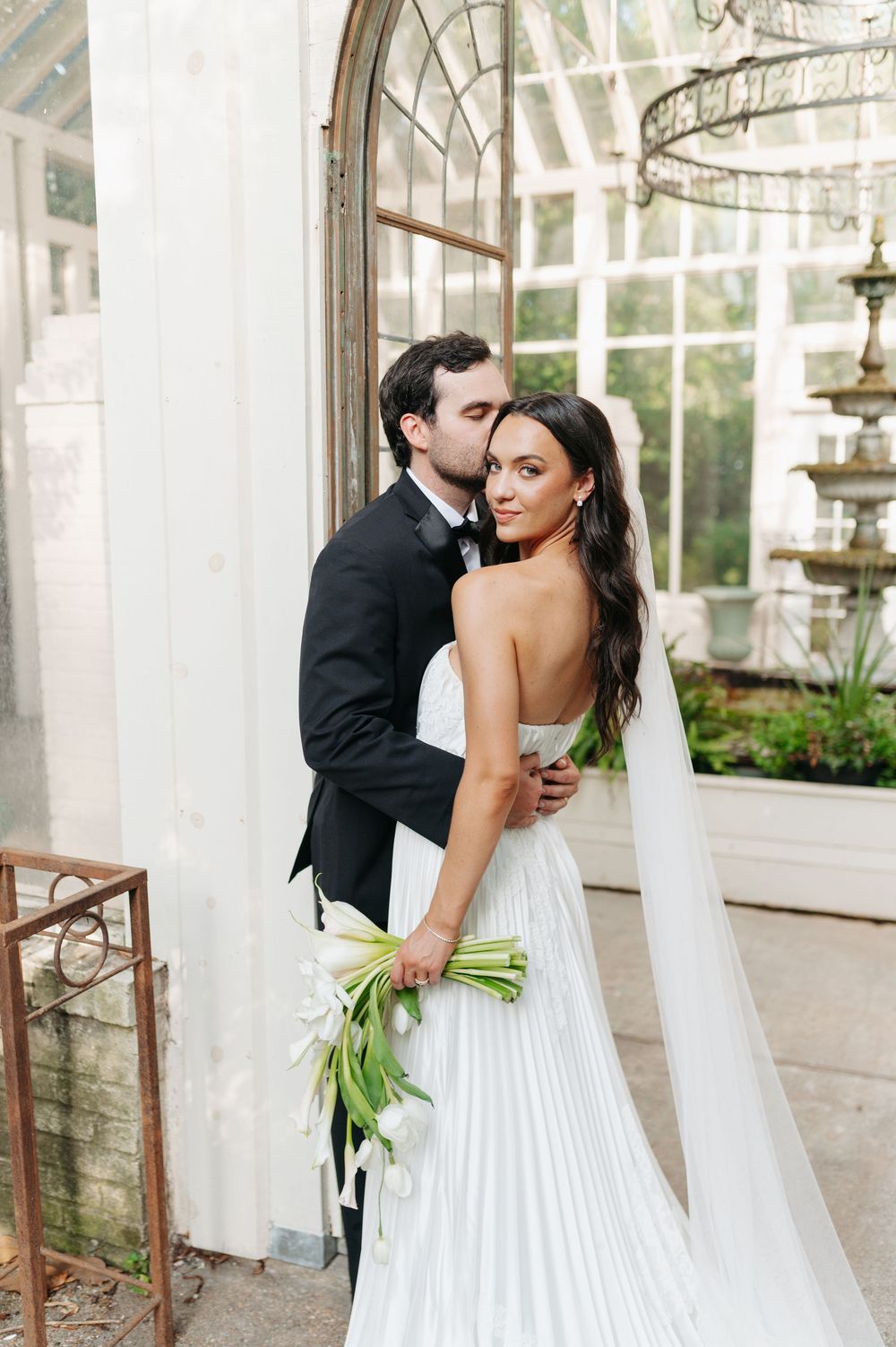 Bride in white pleated gown holds tulips as groom in black tuxedo embraces her outside a glass greenhouse.