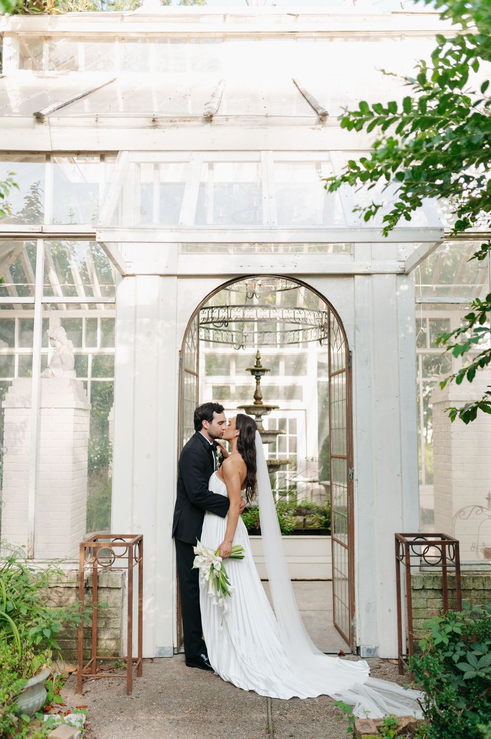 Bride and groom share a romantic kiss in a white greenhouse doorway, bride in flowing gown with long veil.