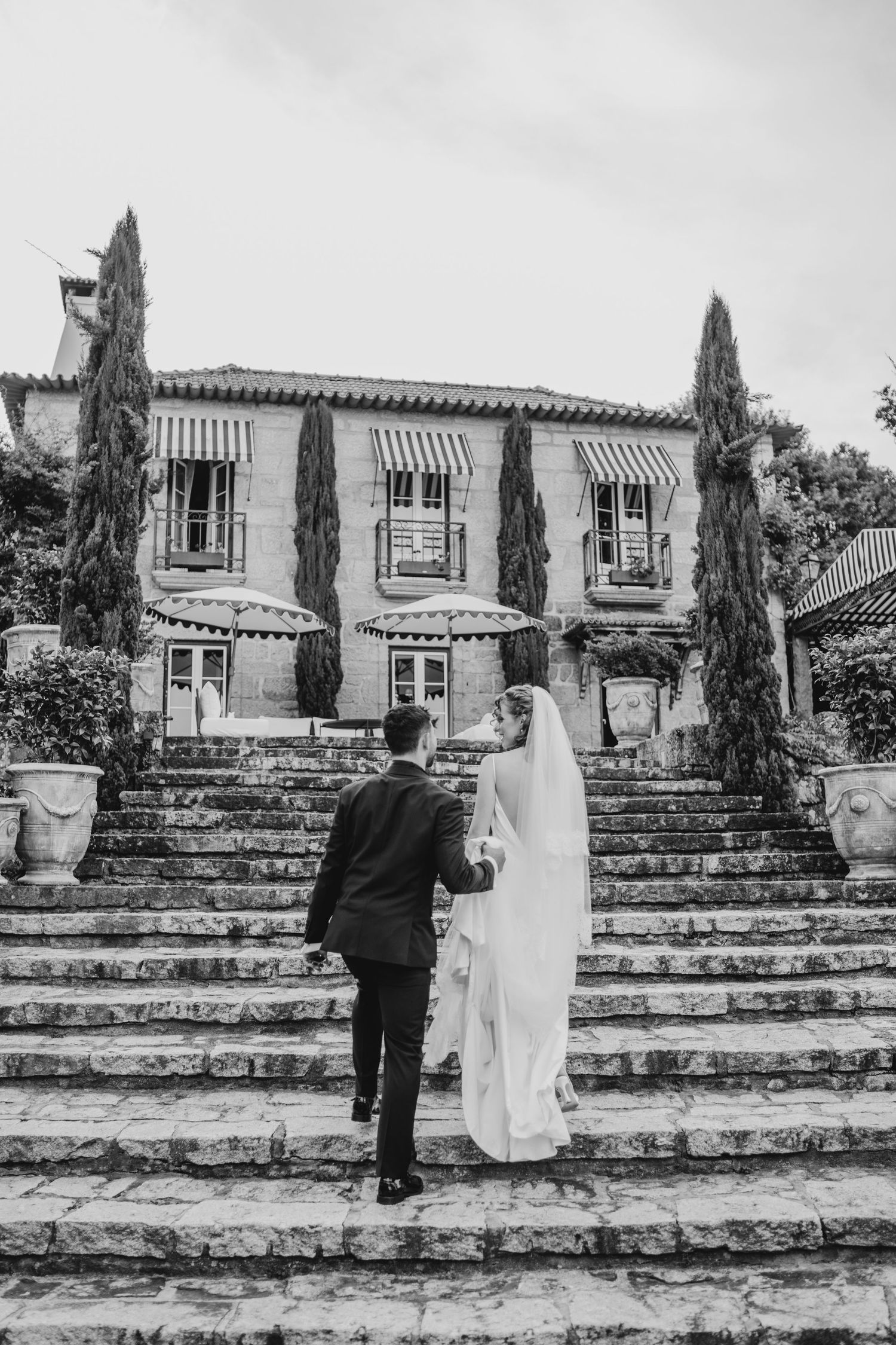 Black and white photo of bride and groom walking up stone steps toward an elegant villa with cypress trees.