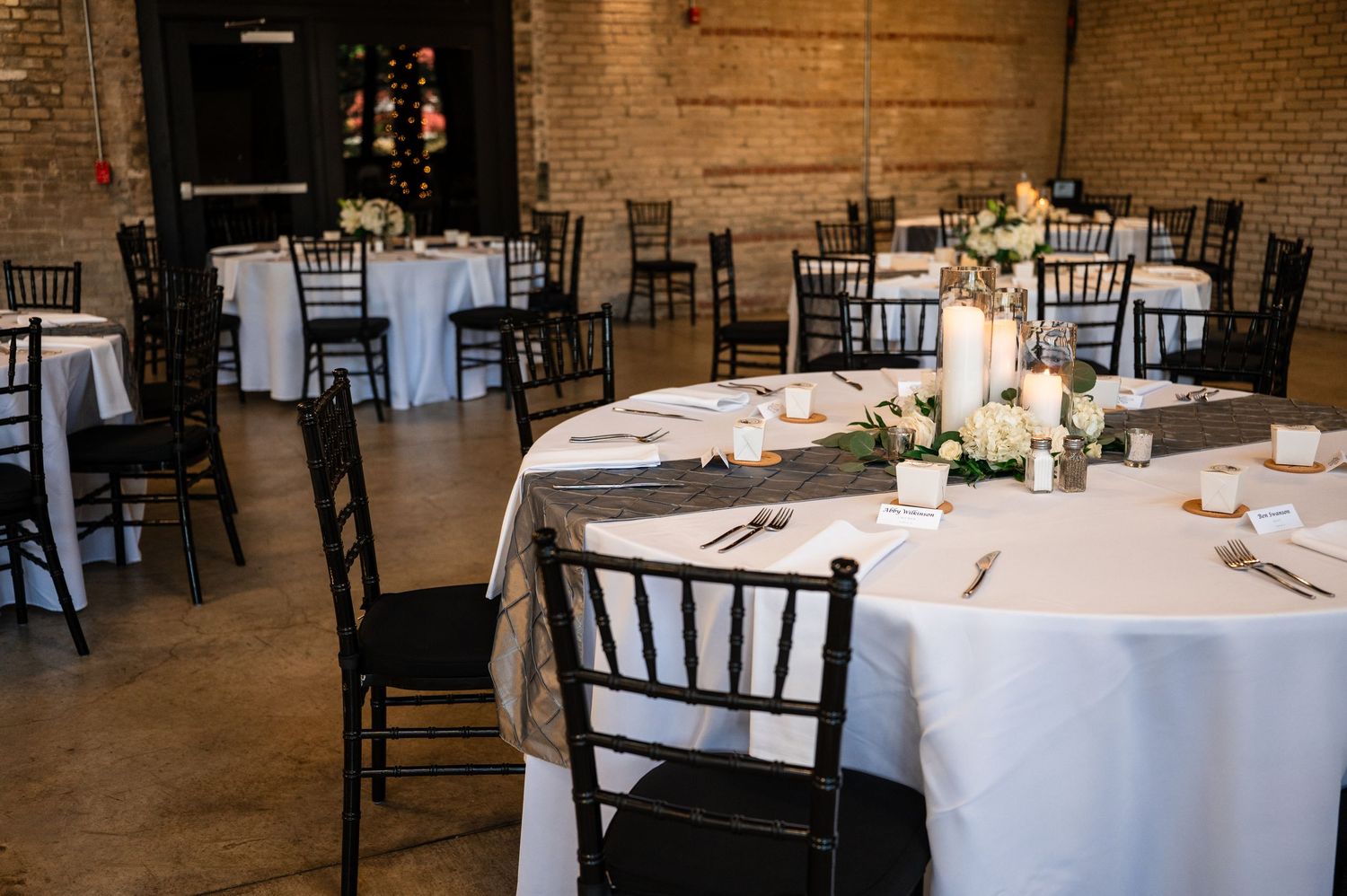 Elegant wedding reception venue with black Chiavari chairs, white tablecloths, and floral centerpieces in a rustic brick-wall setting.