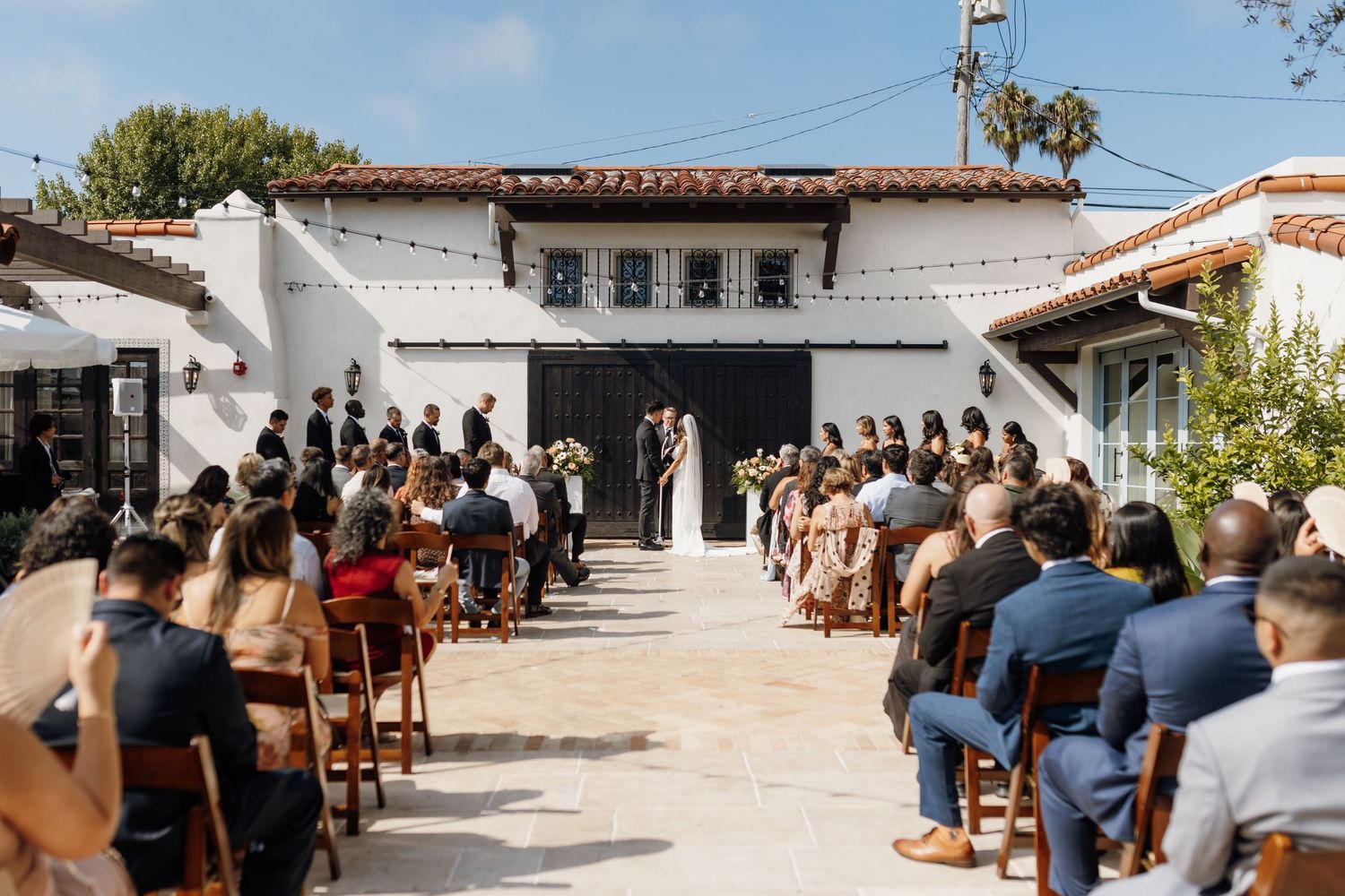 Outdoor wedding ceremony at a Spanish-style venue with guests seated in wooden chairs facing the couple at the altar.