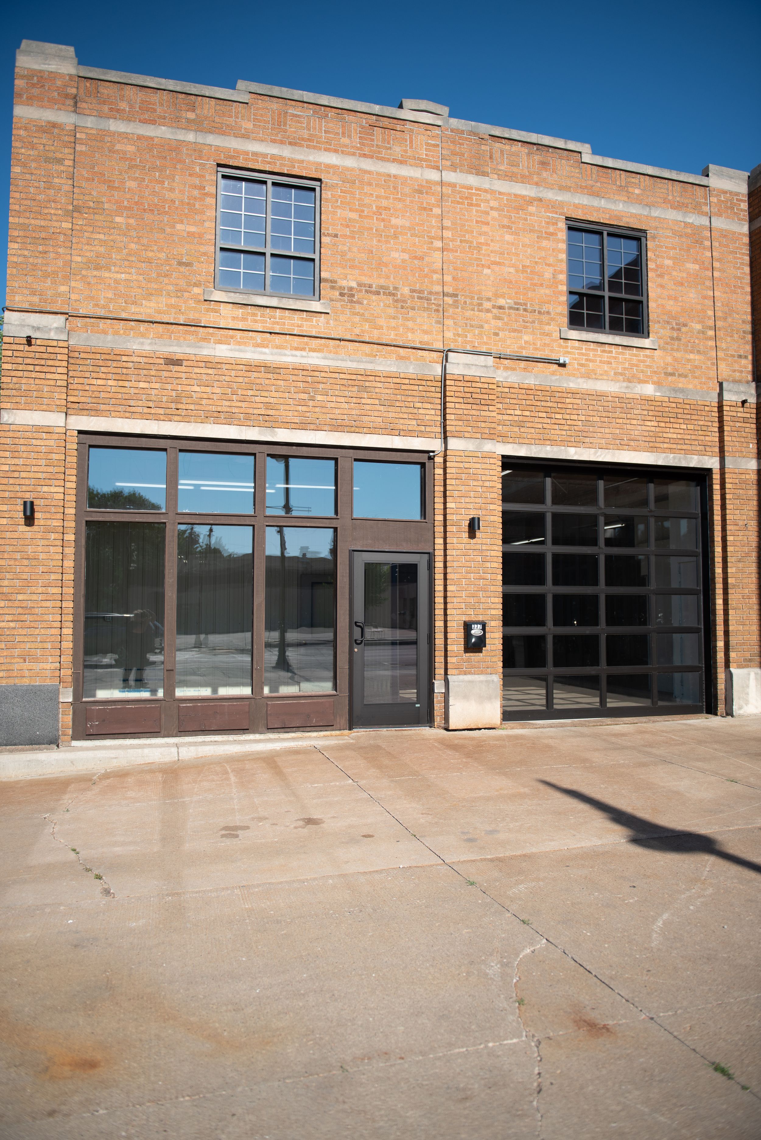 Brick commercial building with large glass doors and garage entrance on concrete lot under blue sky.