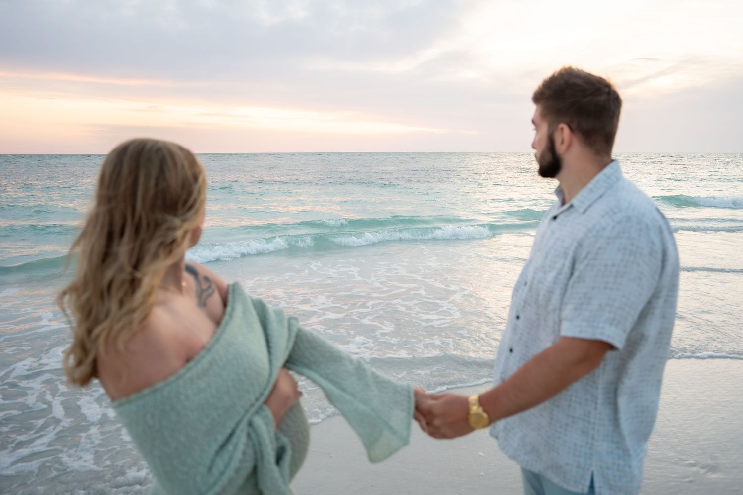 Couple holding hands walking along turquoise ocean shoreline at colorful sunset beach.