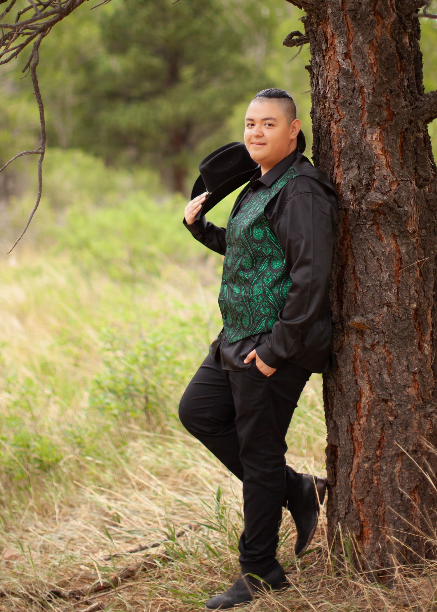 Person in black outfit with green vest leaning against pine tree in natural outdoor setting.