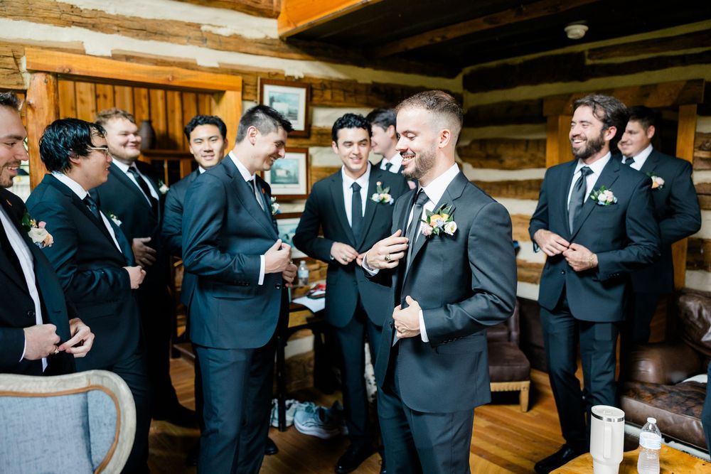 Groom and groomsmen in dark suits with boutonnieres laughing together in a rustic log cabin before the wedding.