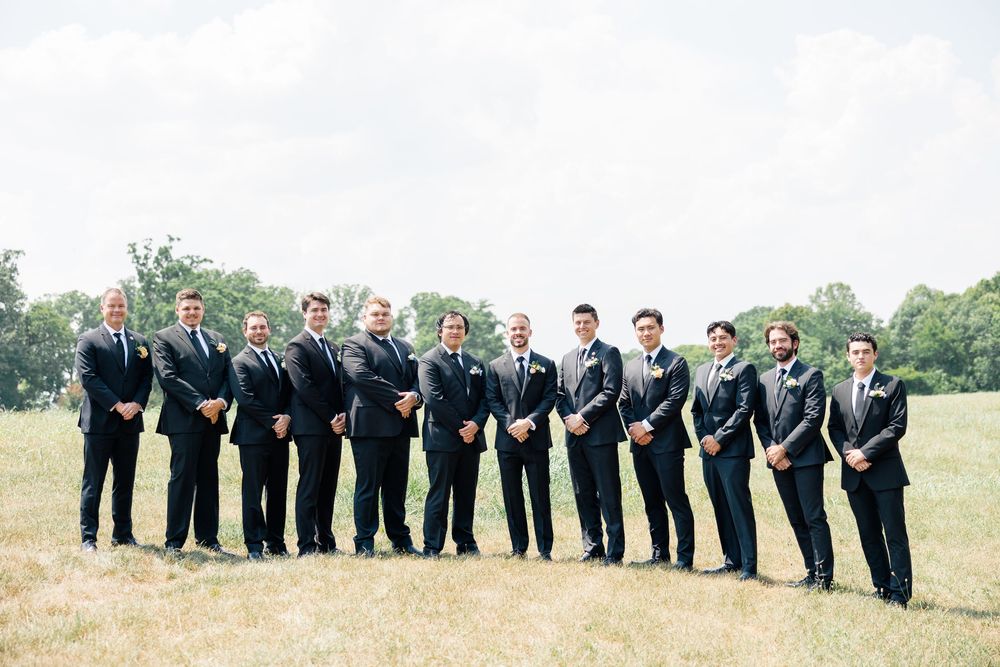 Groom and groomsmen in matching dark suits posing together outdoors on a green field at a wedding.