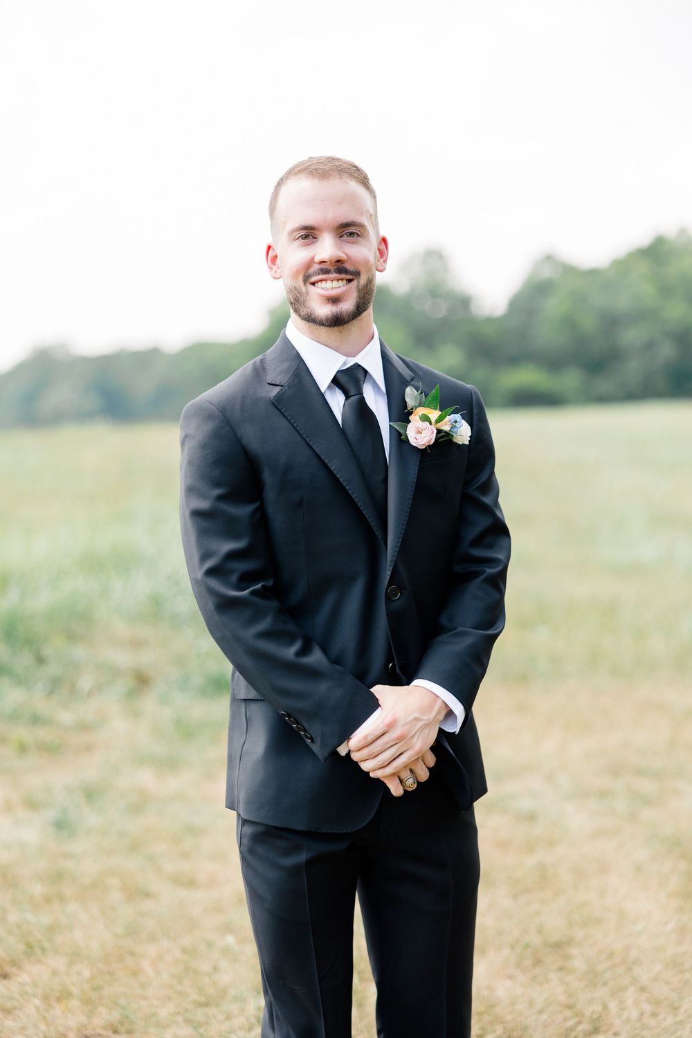 Smiling groom in black suit with floral boutonniere standing in green field on wedding day.