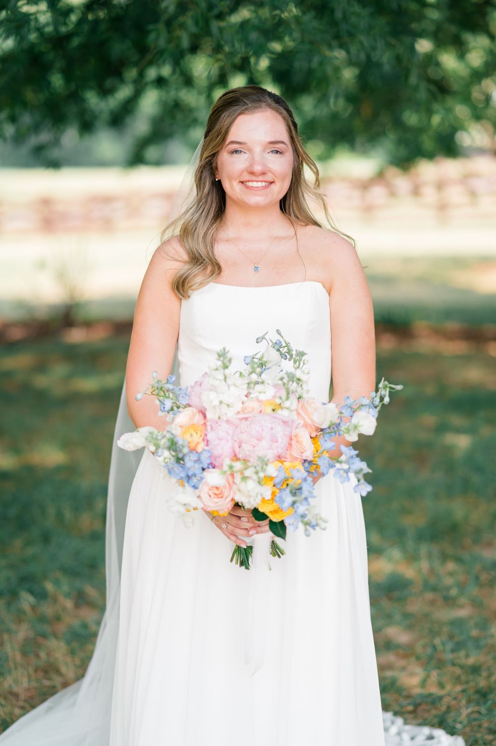 Smiling bride in strapless white gown holding colorful spring bouquet with pink, blue, and yellow flowers outdoors.