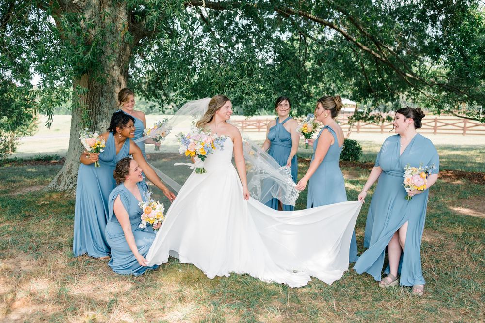 Bride in white gown with bridesmaids in blue dresses holding bouquets under a large tree outdoors.
