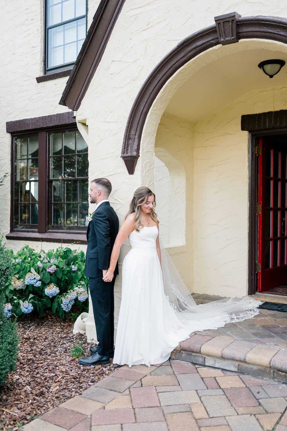 Bride and groom hold hands outside a Tudor-style venue, bride in a flowing white gown with veil, groom in black tux.