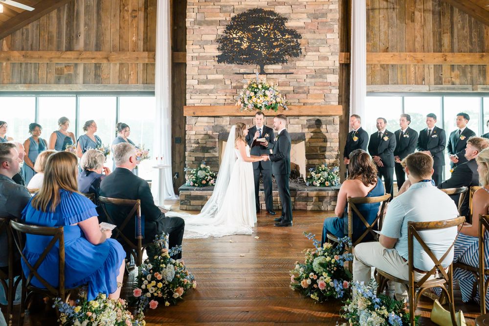 Indoor barn wedding ceremony with couple at floral altar, wood ceiling, stone fireplace, and seated guests in rustic venue.