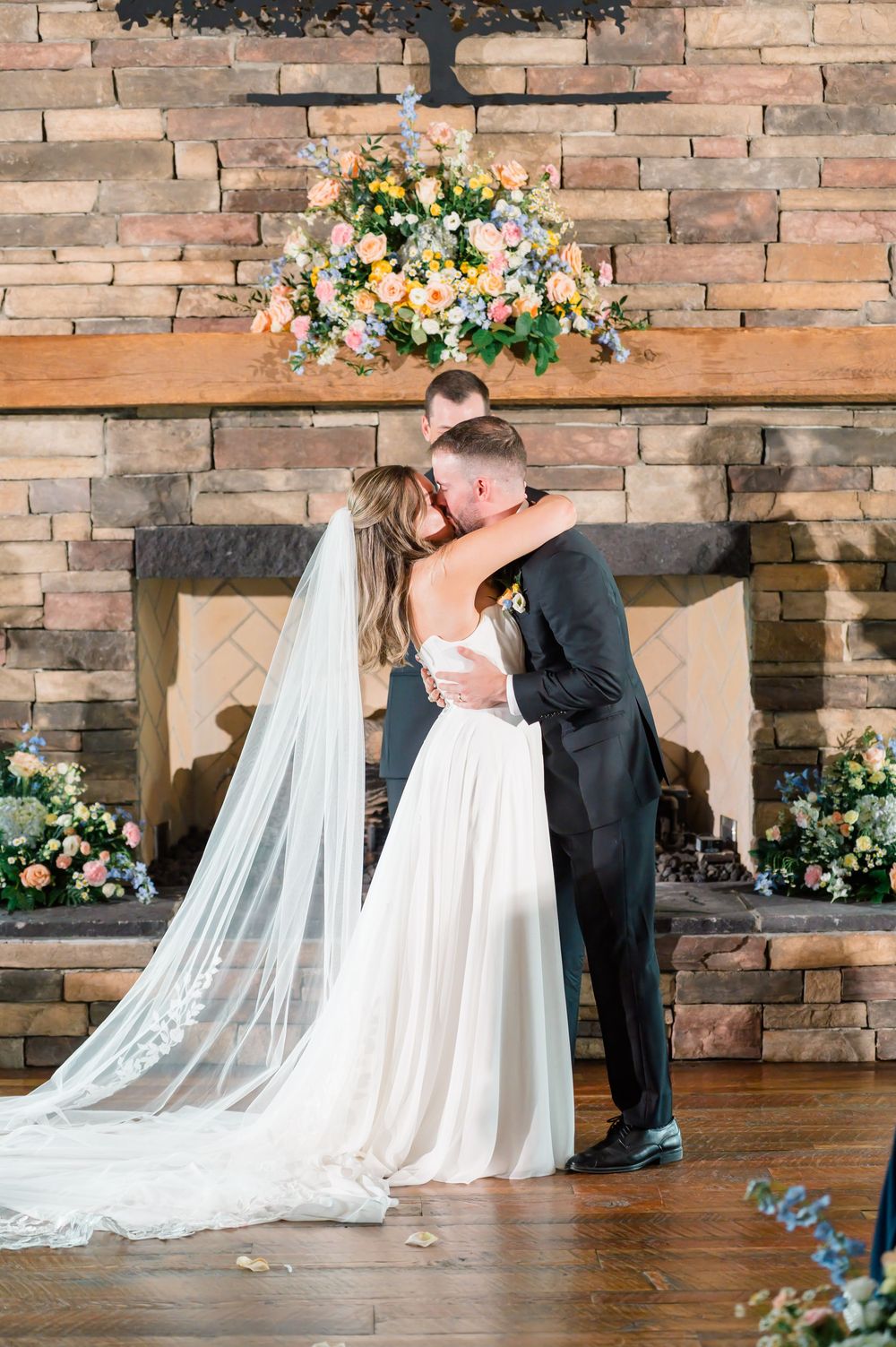 Bride and groom share first kiss at rustic stone fireplace altar adorned with colorful floral arrangements.