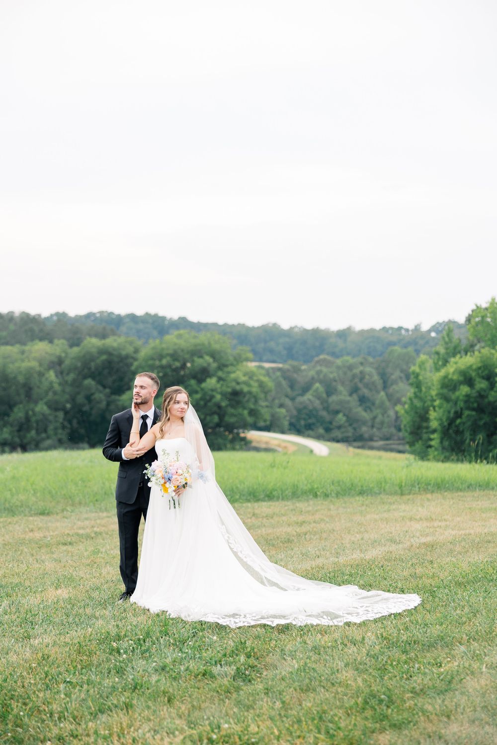 Bride and groom posing in a lush green field, bride wearing a long veil and holding a colorful bouquet.