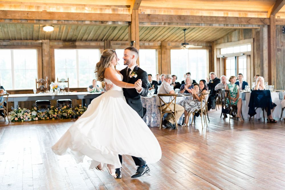 Bride and groom share their first dance at a rustic barn wedding reception with guests seated in background.