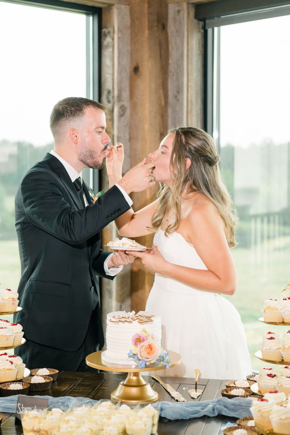 Bride and groom feeding each other wedding cake at a rustic reception with cupcakes displayed on the table.