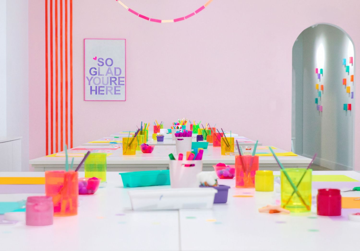 Colorful art studio with floral arrangements, mini easels, paint supplies, and a pink 'So Glad You're Here' sign.