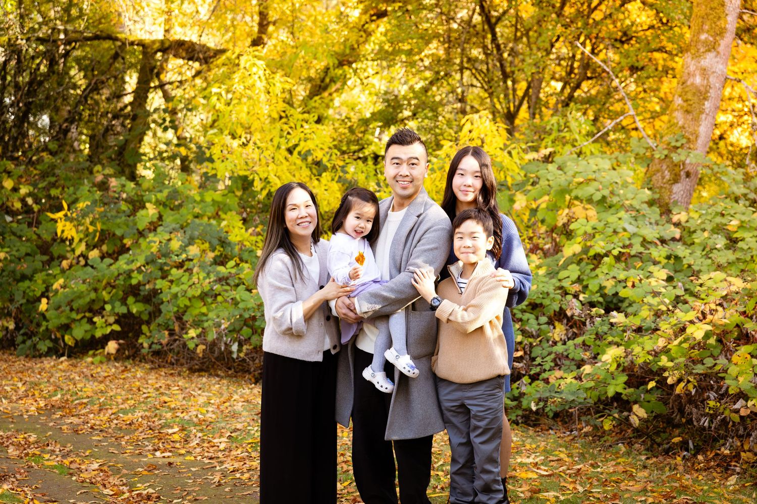 Family of five smiling outdoors among vibrant yellow autumn foliage on a scenic tree-lined path.