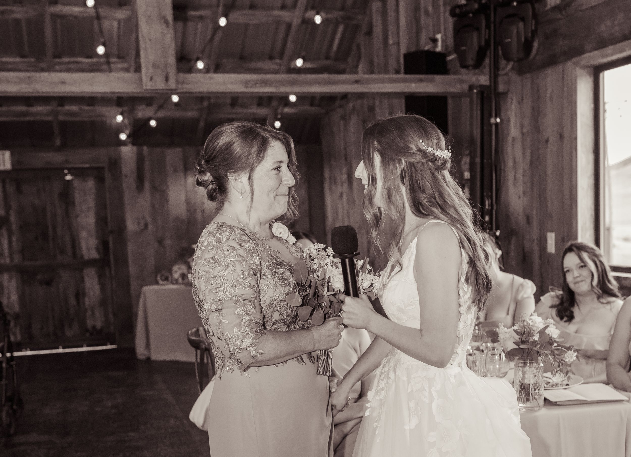 Bride and mother sharing heartfelt moment during wedding reception speech in rustic barn venue.