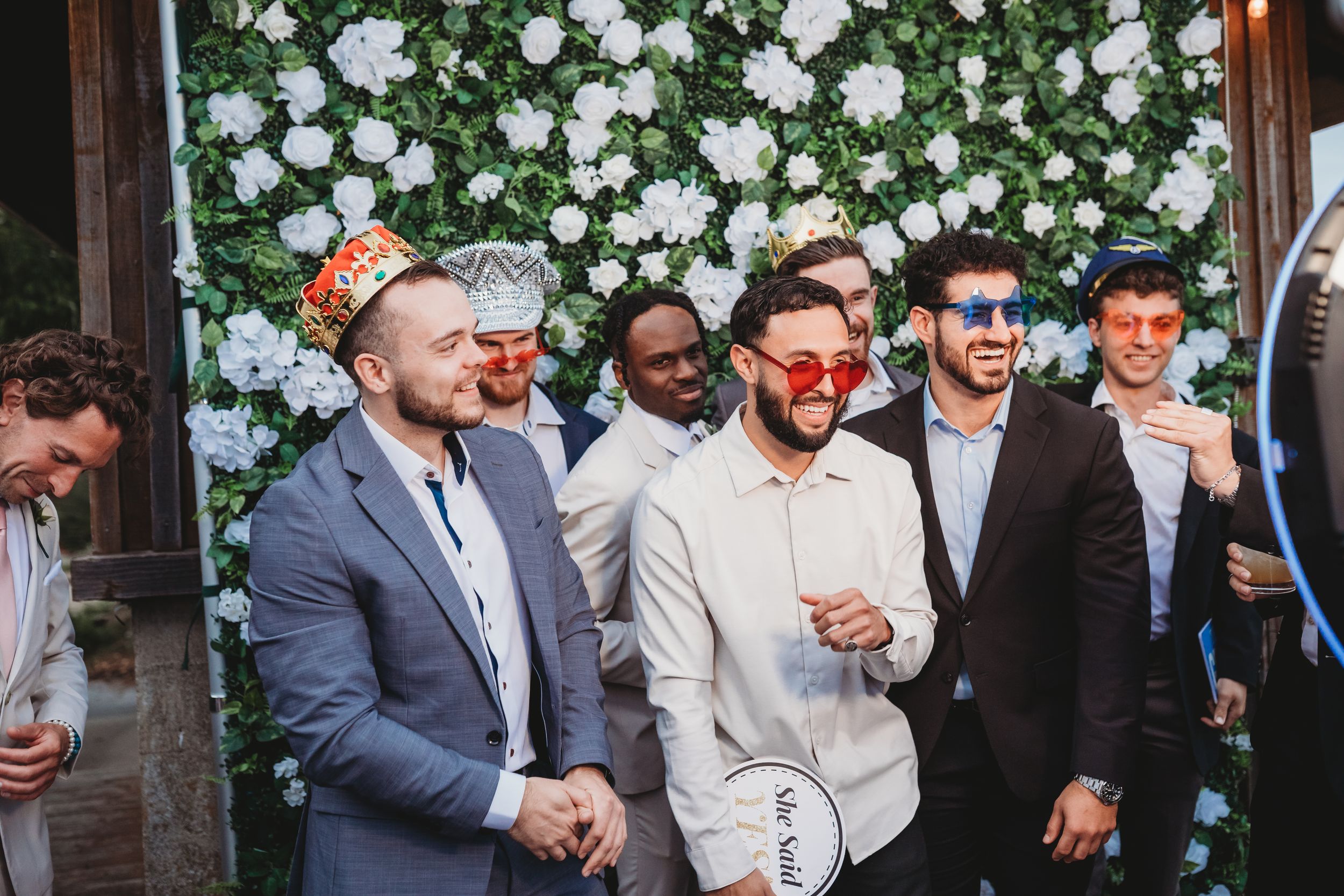 Group of groomsmen wearing fun photo booth props and accessories in front of white flower wall backdrop.