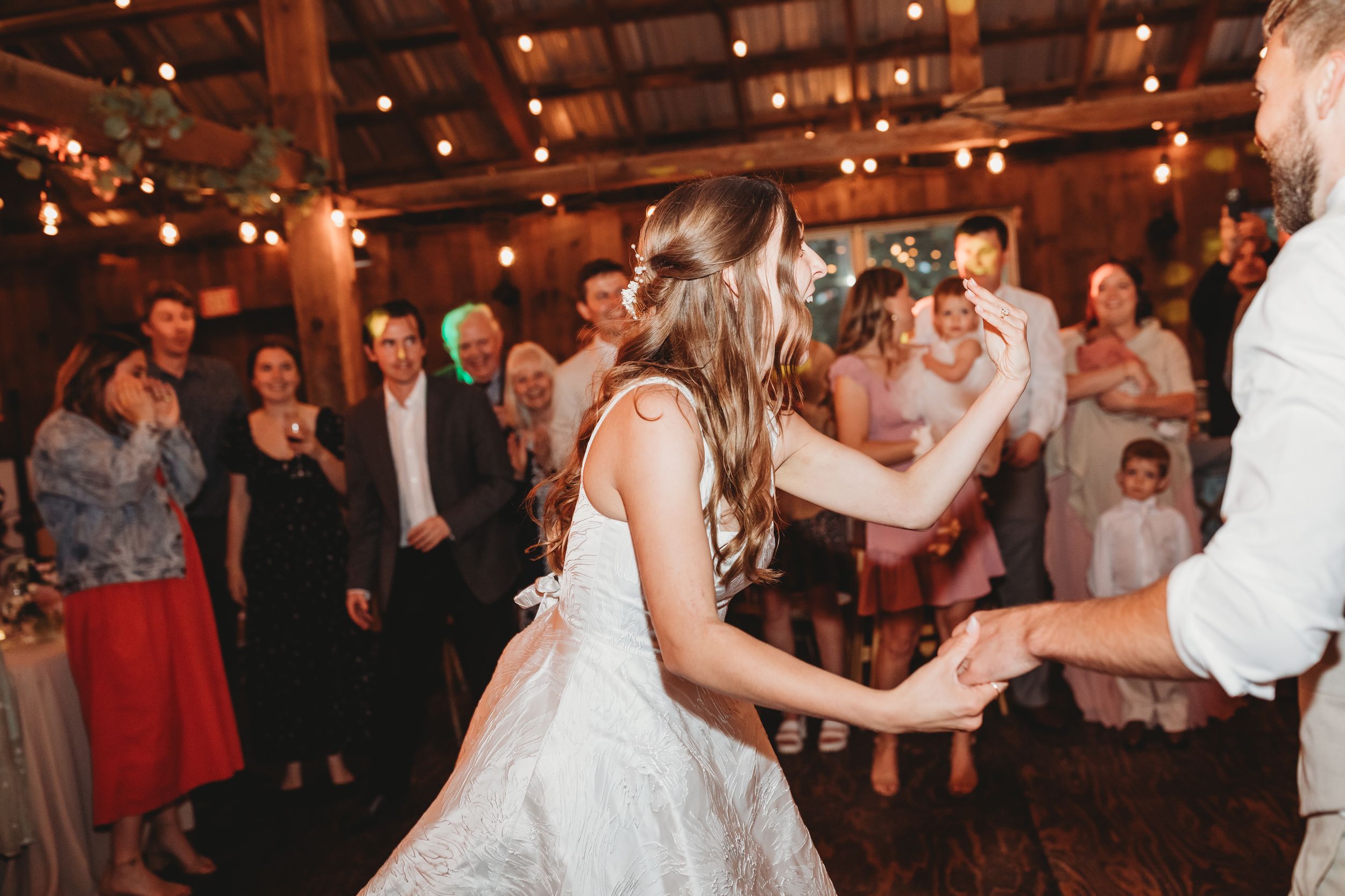 Bride and groom share first dance surrounded by guests under warm string lights at rustic wedding reception.