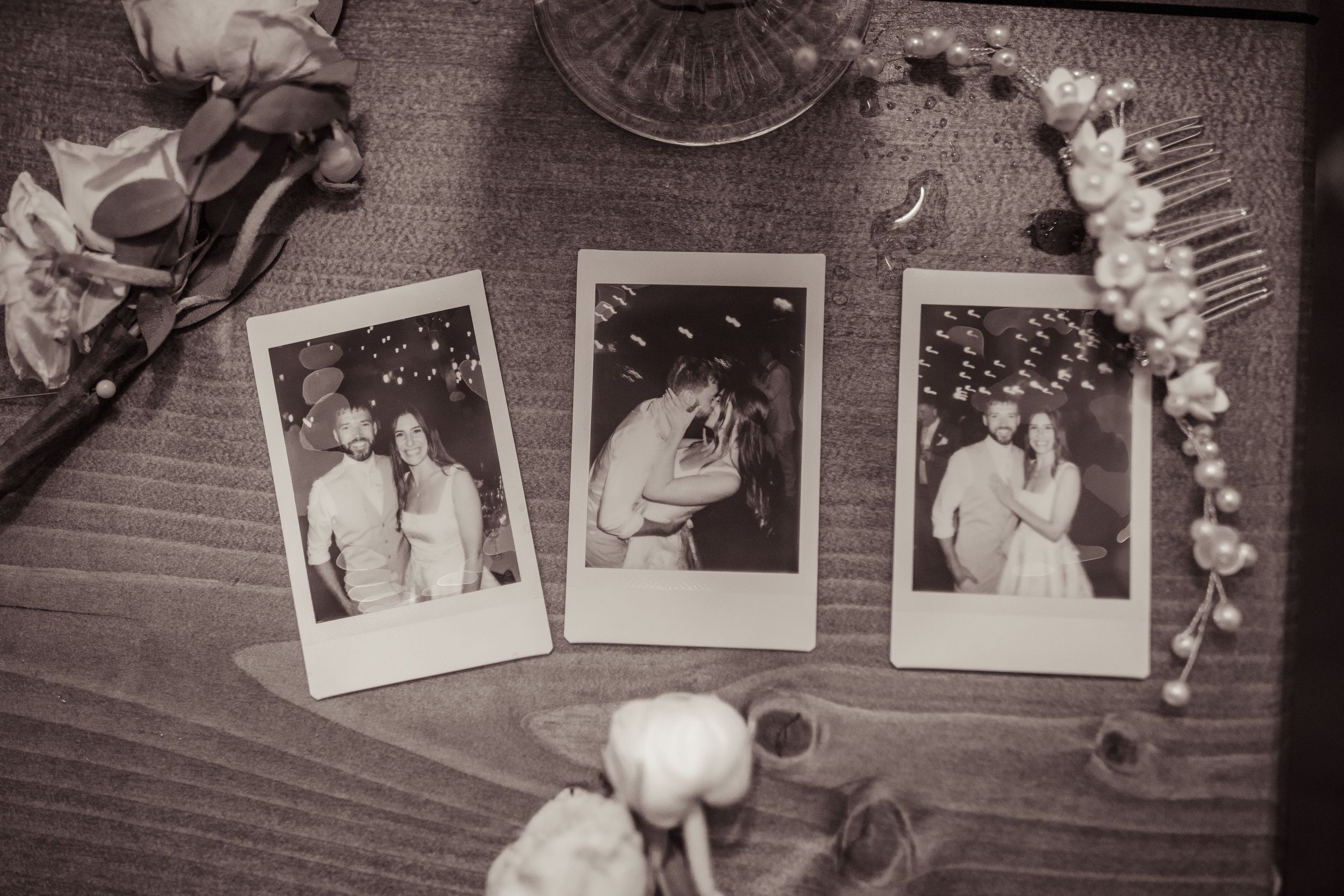 Black and white photo of three Polaroid wedding snapshots on a wooden table surrounded by flowers and a pearl hair comb.