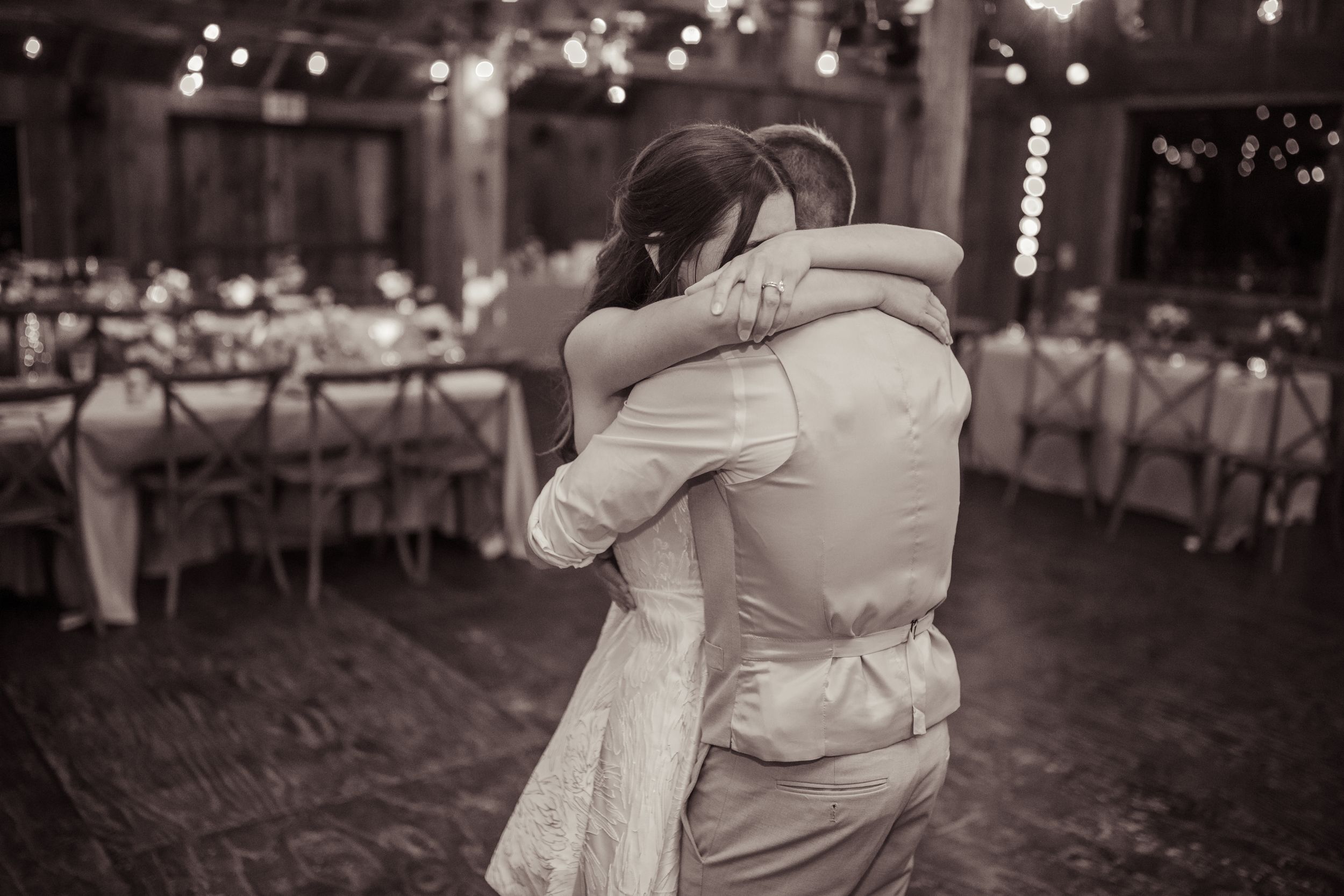 Bride and groom share tender embrace during first dance in elegant rustic reception hall, black and white photo.