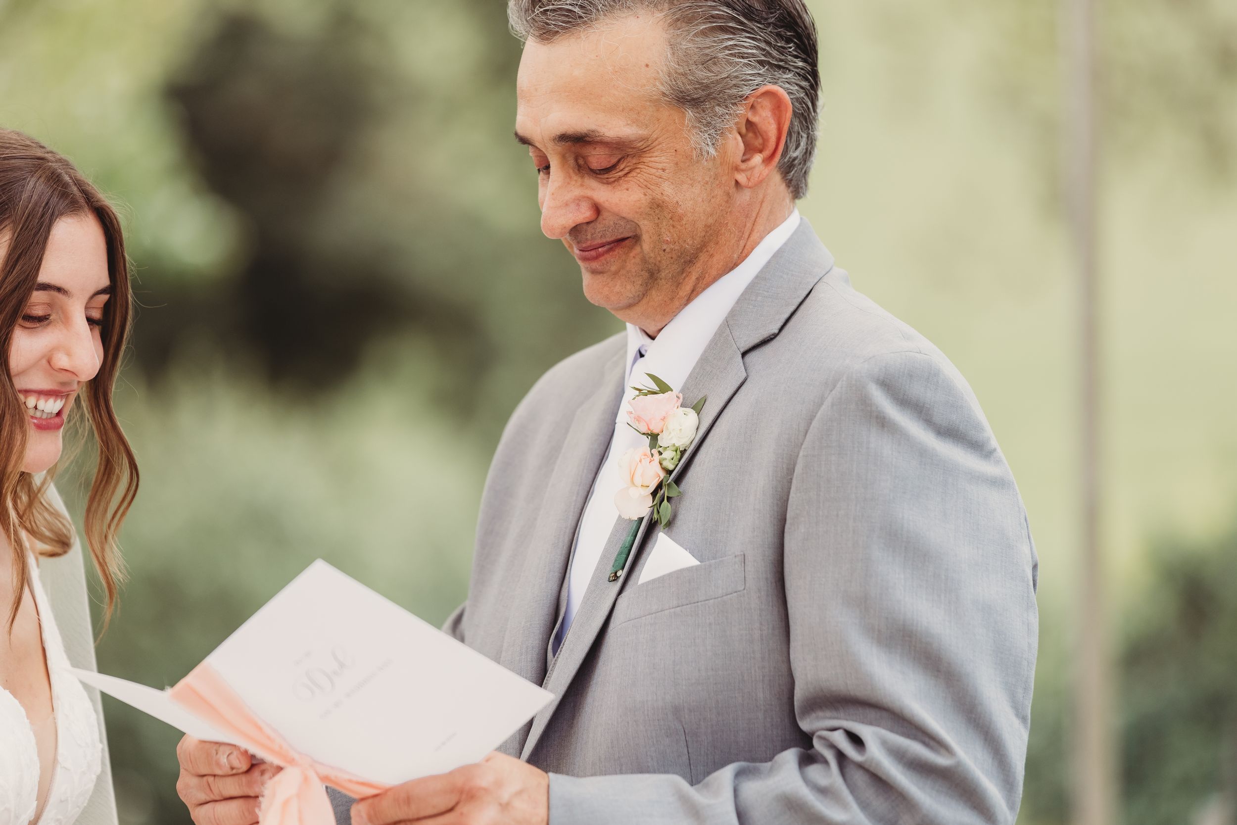 Groom in gray suit reading wedding vows at outdoor ceremony, smiling bride beside him.