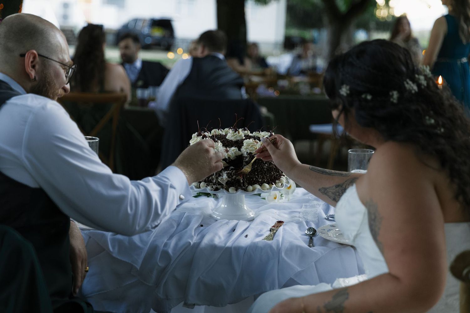 Bride and groom cutting their wedding cake at an outdoor evening reception with guests seated in background.