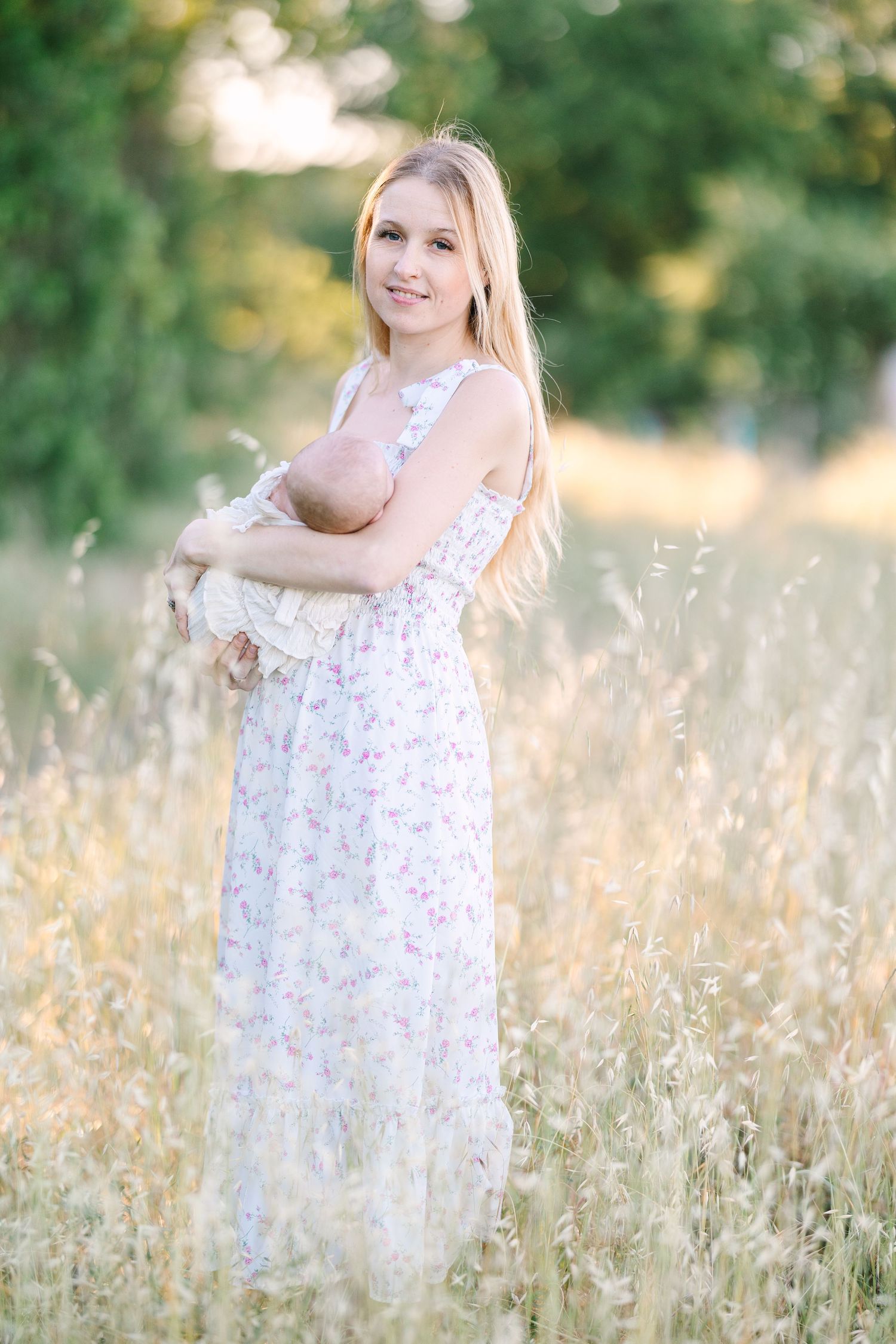 Blonde woman in floral dress holding newborn baby in golden meadow with green trees in background.