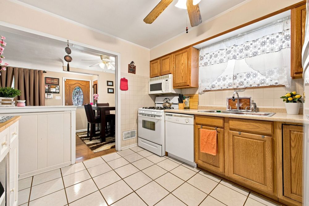 Bright kitchen with oak cabinets, white appliances, tile floors, and a window with lace curtains overlooking a dining area.